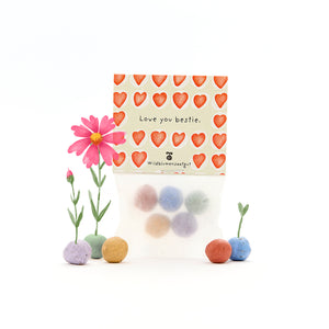 Tüte mit Wildblumensamen, dekoriert mit roten Herzen und der Aufschrift "Love you bestie.", umgeben von bunten Samenbällchen und kleinen blühenden Blumen.