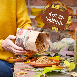 Eine Person in gelbem Pullover füllt Igel-Futter aus einer Papiertüte in eine flache Schale, umgeben von Herbstlaub; daneben ein Schild mit der Aufschrift "Ideal zum Nachfüllen".