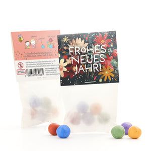 Zwei Tüten mit bunten Saatkugeln, eine mit blumigem "Frohes neues Jahr!"-Aufdruck und eine mit Beschreibung und Zeichnungen, einige Kugeln liegen davor.