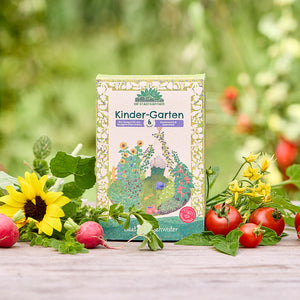 Eine Packung „Kinder-Garten“ Bio-Saatgut steht auf einem Holztisch, umgeben von Sonnenblume, Radieschen, Tomaten und frischem Grün im Garten.