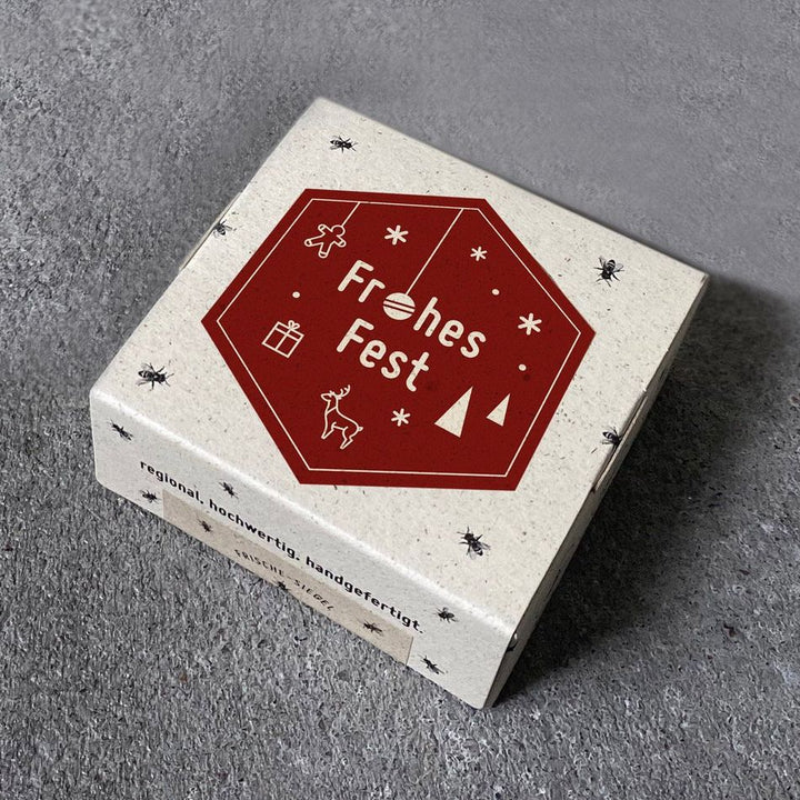 Geschenkbox mit der Aufschrift "Frohes Fest" und weihnachtlichen Symbolen auf grauem Untergrund.