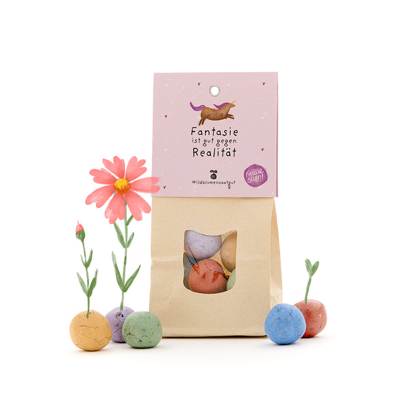 Papiertüte mit bunten Wildblumensamenkugeln, einige mit kleinen Blumen, und Etikett mit Einhorn und dem Text „Fantasie ist gut gegen Realität“.