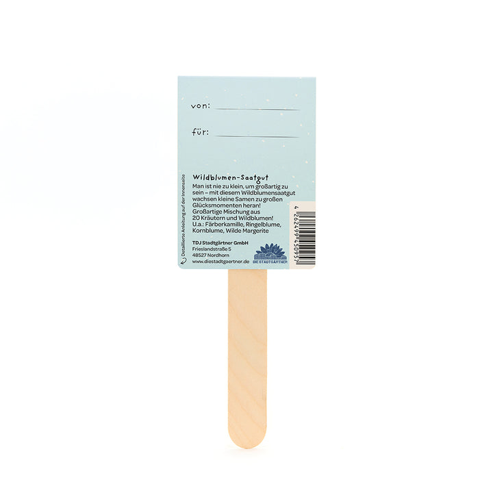 Rückseite eines Wildblumensaatgut-Sticks mit Beschreibungstext, Platz für "von" und "für"-Eintrag, sowie Produkt- und Herstellerinformationen auf hellblauem Hintergrund.