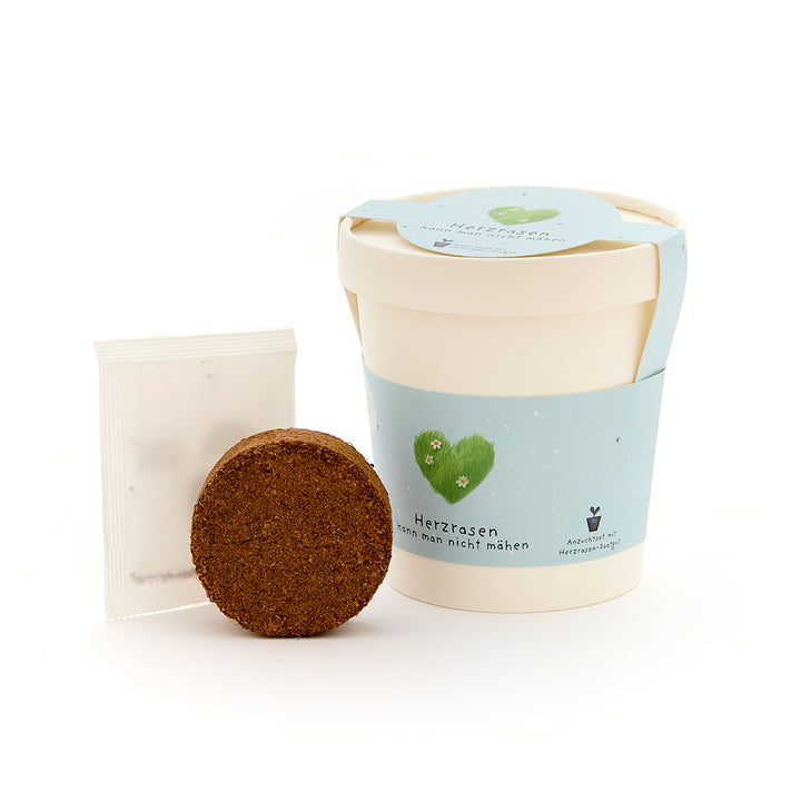 Anzuchtset mit weißem Becher, Erde und Samenpäckchen, mit blauem Etikett "Herzrasen".