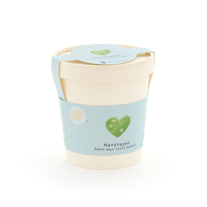 Beiger Pflanztopf mit hellblauem Etikett, grünem Herz und der Aufschrift „Herzrasen kann man nicht mähen“.