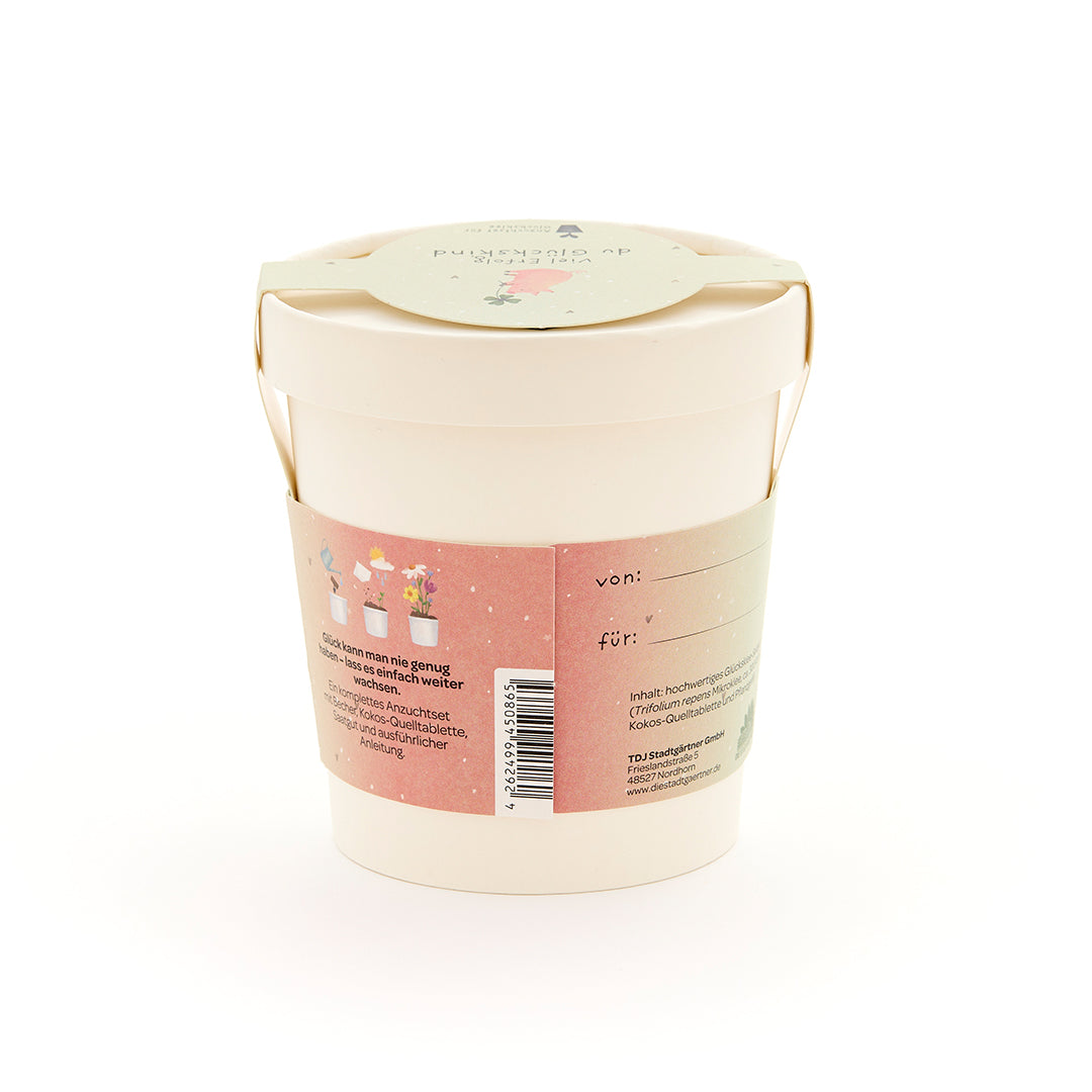 Beiger Blumentopf mit Henkel, rosa Etikett mit Pflanzillustrationen und Text auf Deutsch, Geschenkanhänger auf dem Deckel.