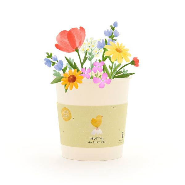 Illustrierter Blumentopf mit bunten Blumen und gelbem Etikett, auf dem ein Küken und der Text "Hurra, du bist da!" zu sehen sind.