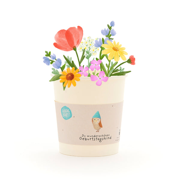 Ein Blumentopf mit bunten Blumen und einer Banderole mit der Aufschrift „Du wunderschönes Geburtstagskind“.