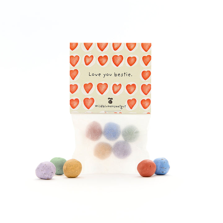 Tüte mit bunten Samenbällen, darauf eine Karte mit roten Herzen und dem Text "Love you bestie. Wildblumensaatgut".
