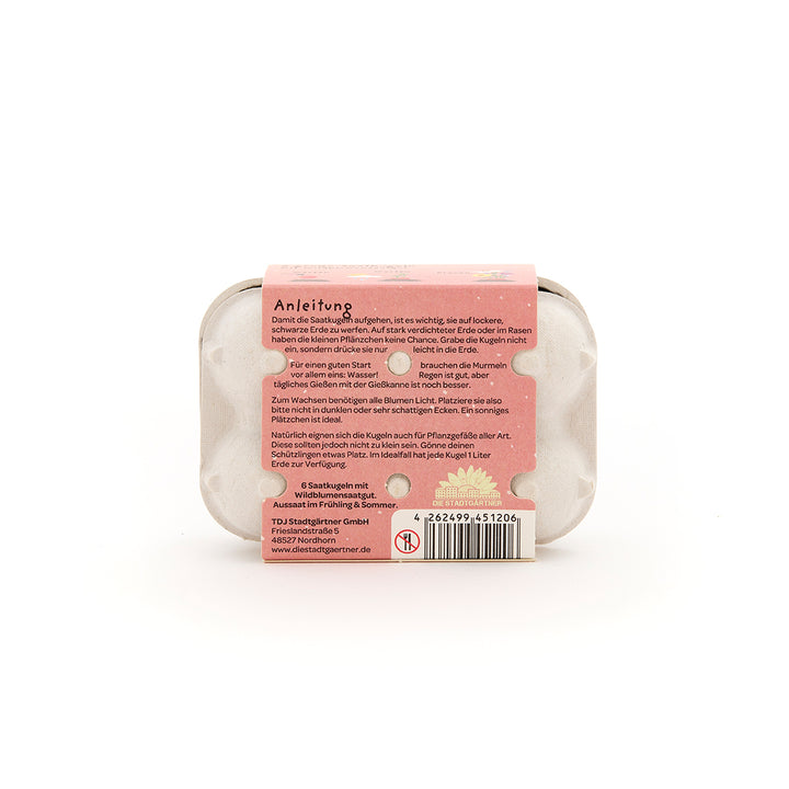 Eine kleine Kartonverpackung mit rosa Anleitungsetikett und Samenbällchen, Rückseite mit Pflegehinweisen auf Deutsch.