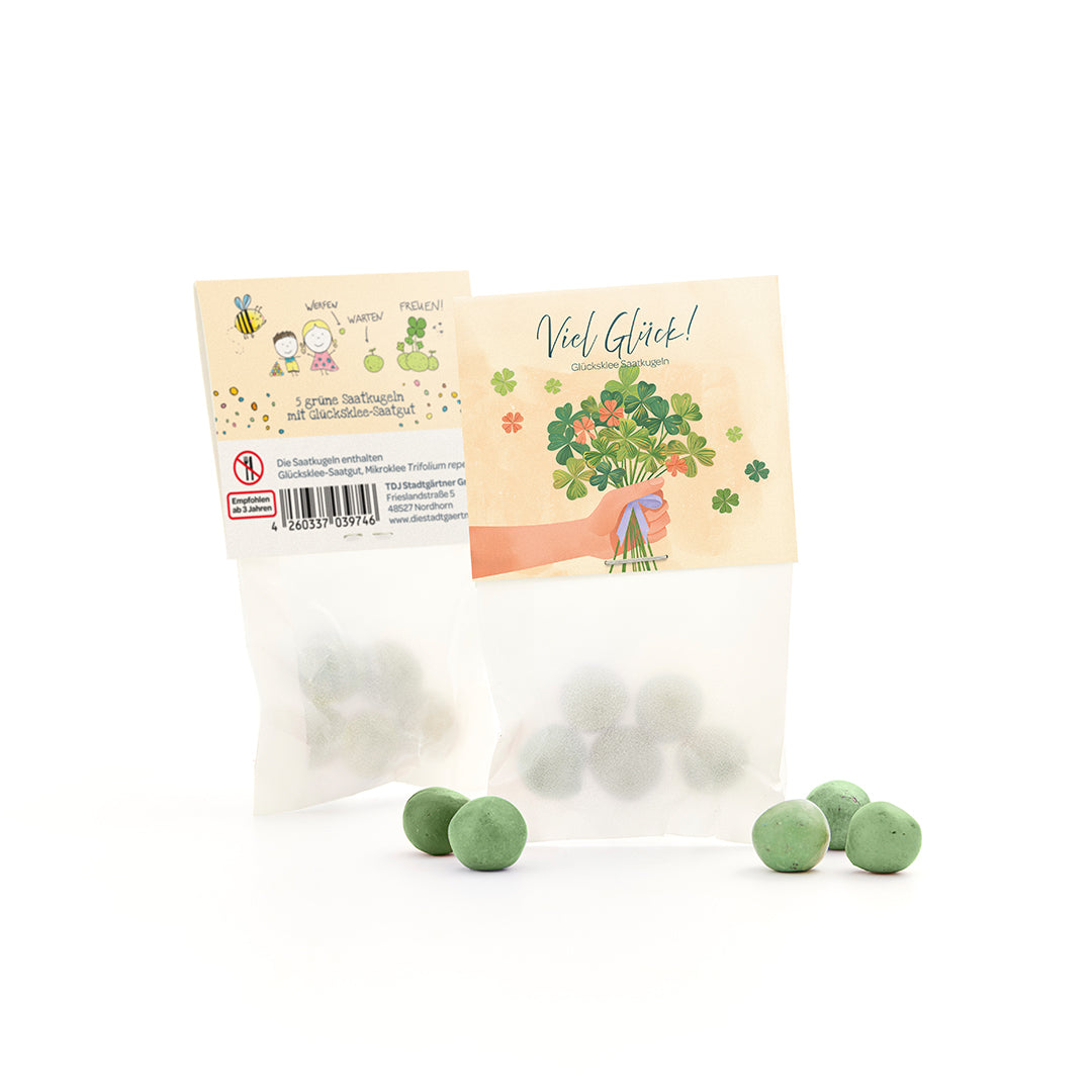 Tüte mit grünen Saatkugeln und Glücksklee-Samen, daneben mehrere lose grüne Kugeln, auf der Verpackung Glückwunschmotiv und Kleeillustration.