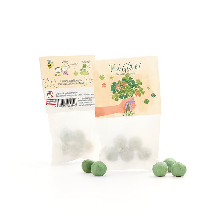 Tüte mit grünen Saatkugeln und Glücksklee-Samen, daneben mehrere lose grüne Kugeln, auf der Verpackung Glückwunschmotiv und Kleeillustration.