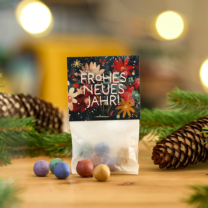 Tüte mit bunten Kugeln und einer Karte mit der Aufschrift "Frohes neues Jahr!" auf einem Holztisch, umgeben von Tannenzweigen und Tannenzapfen.