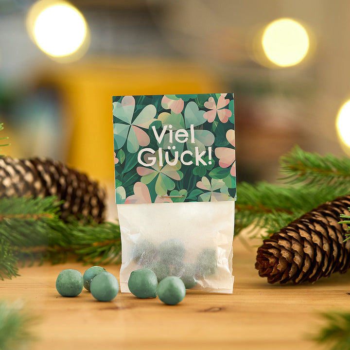 Tütchen mit grünen Kugeln und der Aufschrift "Viel Glück!" vor weihnachtlicher Dekoration mit Tannenzweigen und Tannenzapfen.