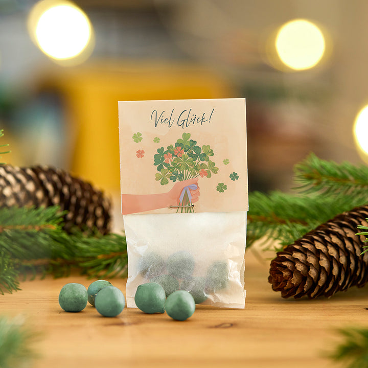 Tüte mit grünlichen Kugeln, dekoriert mit einer Karte, auf der „Viel Glück!“ und ein Kleeblattstrauß abgebildet sind, umgeben von Tannenzweigen und Zapfen.