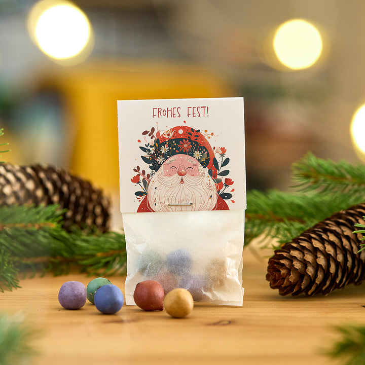 Tüte mit bunten Kugeln, Weihnachtskarte mit Santa-Motiv und Aufschrift „Frohes Fest!“, Tannenzapfen und Zweige im Hintergrund.