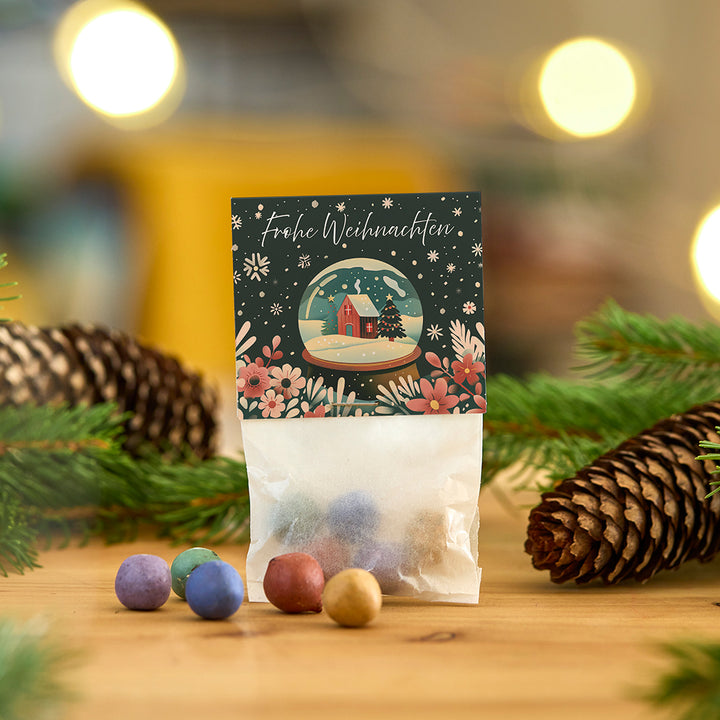 Tüte mit bunten Kugeln und weihnachtlicher Karte mit der Aufschrift "Frohe Weihnachten" vor weihnachtlicher Dekoration mit Tannenzapfen und Zweigen.