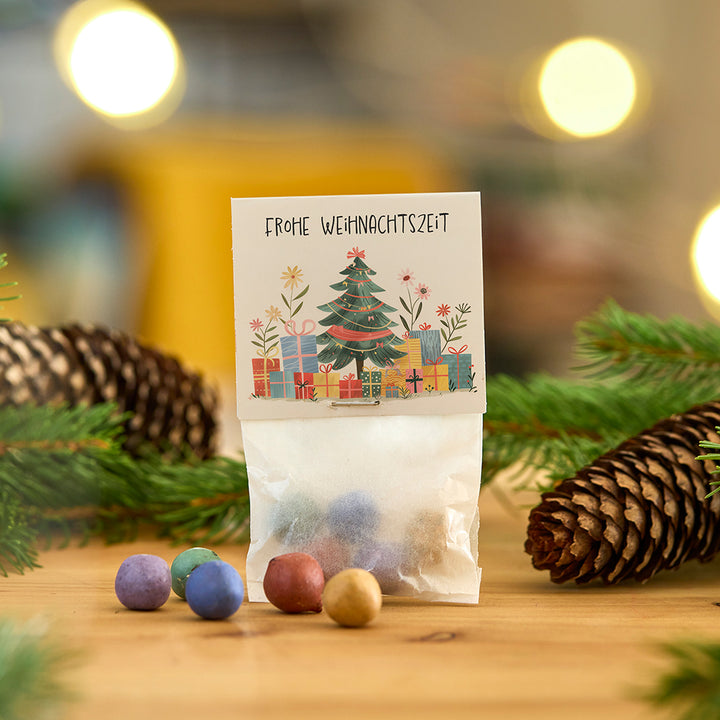 Eine kleine Tüte mit bunten Kugeln, dekoriert mit einer Weihnachtskarte mit der Aufschrift "Frohe Weihnachtszeit", umgeben von Tannenzweigen und einem Tannenzapfen.