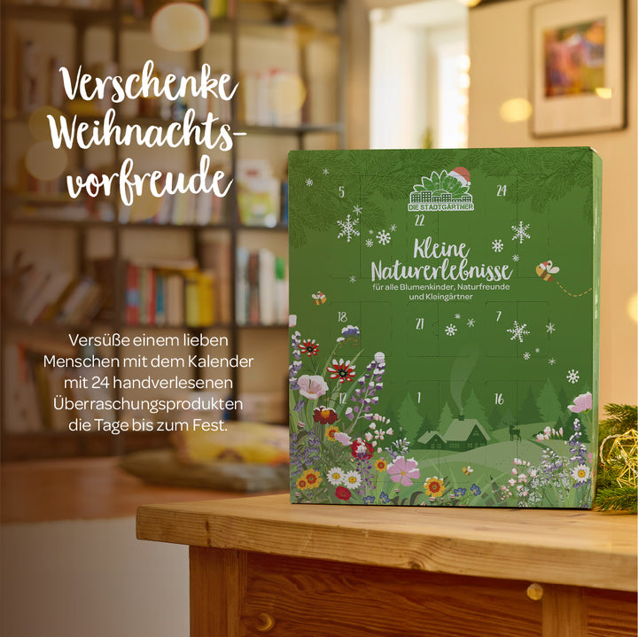 Grüner Adventskalender mit Blumenmotiv steht auf einem Holztisch vor einem Bücherregal, daneben steht der Text "Verschenke Weihnachtsvorfreude".