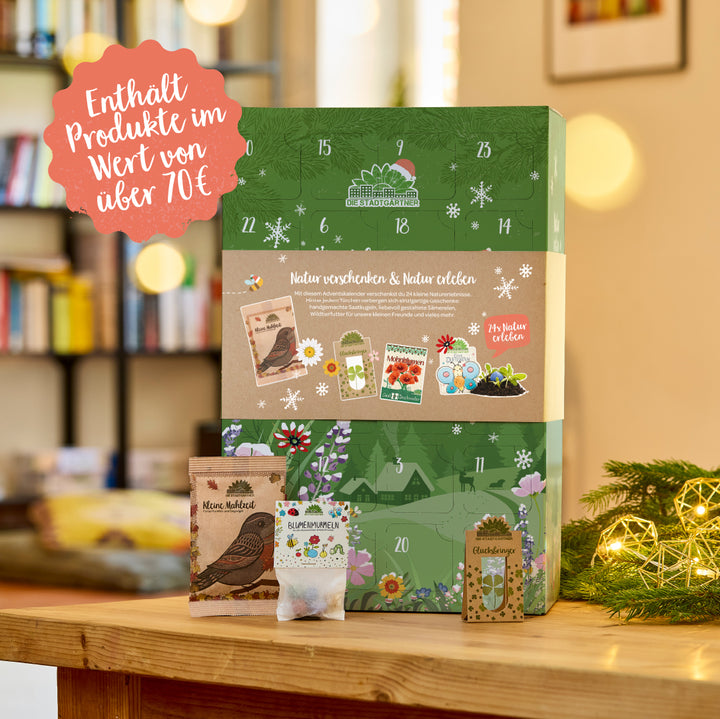Adventskalender mit grünem, winterlichem Design, steht auf einem Holztisch vor einem unscharfen Bücherregal, mit Dekoration und kleinen Tüten für Samen und Blumenkugeln im Vordergrund; Text auf dem Kalender und einer roten Blase weist auf Produkte im Wert von über 70 Euro hin.