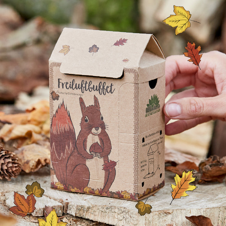 Pappkarton mit Eichhörnchen-Illustration und der Aufschrift "Freiluftbuffet", umgeben von herbstlichen Blättern, wird von einer Hand gehalten.