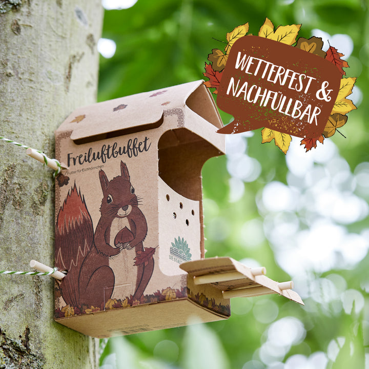 Papp-Futterstation mit Eichhörnchen-Motiv an einem Baum befestigt, mit der Aufschrift „Freiluftbuffet“ und einem Schild „Wetterfest & nachfüllbar“.