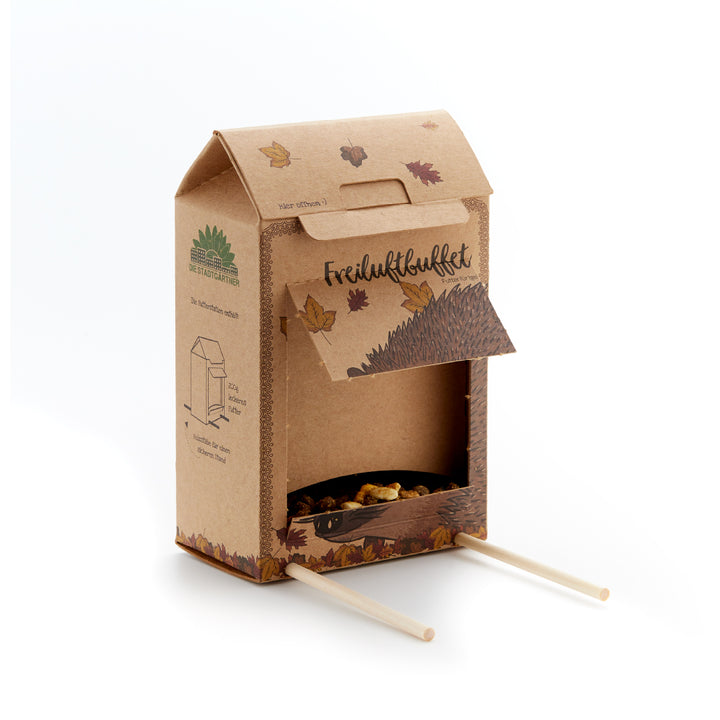 Pappkarton mit geöffneter Klappe, Herbstblättern, Igel-Illustration und zwei Holzstäben, dient als Futterstation für Igel.