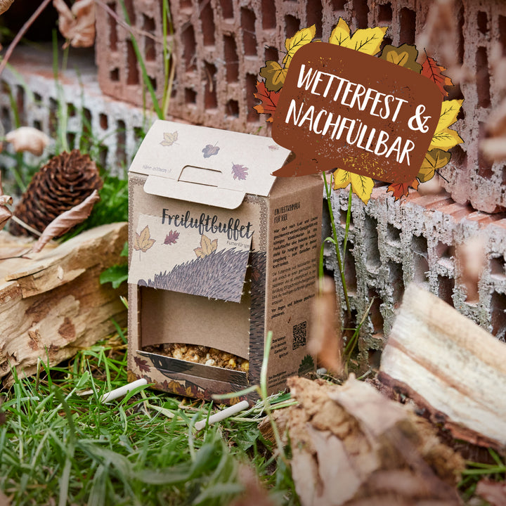Karton mit der Aufschrift "Freiluftbuffet" als wetterfestes und nachfüllbares Futterhaus für Tiere steht im Gras neben Holz und Ziegelsteinen.