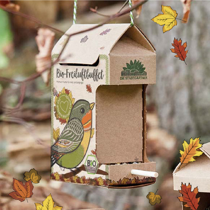 Karton-Vogelfutterhaus mit herbstlichen Blättern, buntem Vogelmotiv und der Aufschrift „Bio-Freiluftbuffet“ an einem Ast hängend.