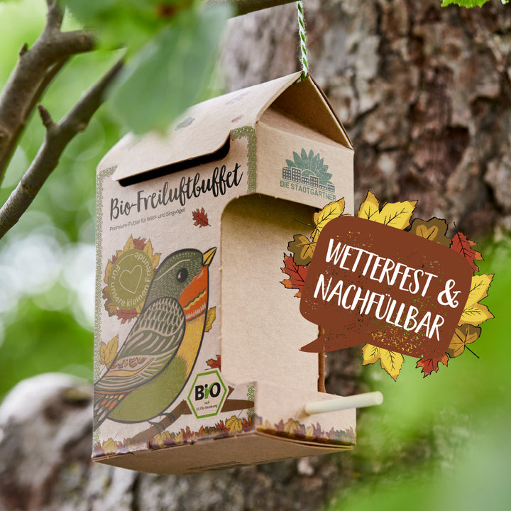 Pappfutterhaus mit Vogelillustration und Aufschrift „Bio-Freiluftbuffet“ hängt an einem Baum, daneben Text „Wetterfest & nachfüllbar“.
