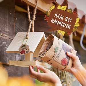 Eine Person füllt ein hölzernes Vogelhaus mit Vogelfutter aus einer Papiertüte; im Bild steht „Ideal zum Nachfüllen“.