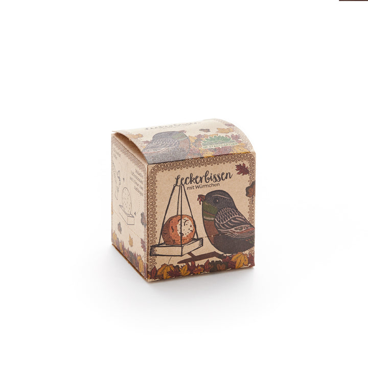 Kartonverpackung mit einer Illustration eines Vogels und eines Meisenknödels, Aufschrift "Leckerbissen mit Würmchen", herbstlich dekoriert.