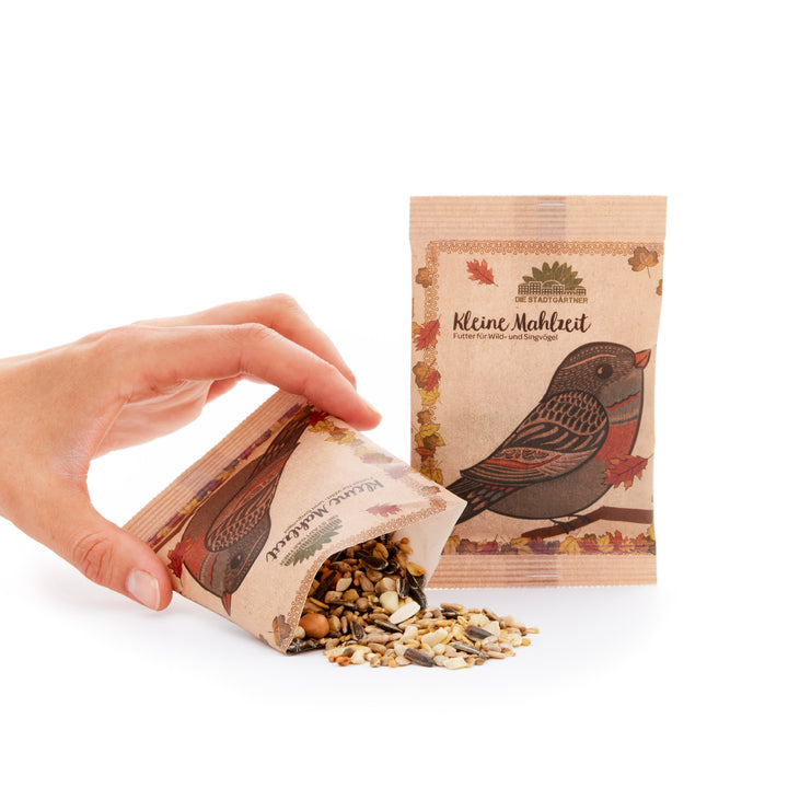 Hand schüttet Vogelfutter aus einer braunen Papiertüte mit Vogelmotiv und der Aufschrift "Kleine Mahlzeit" neben einer stehenden, ungeöffneten Tüte.