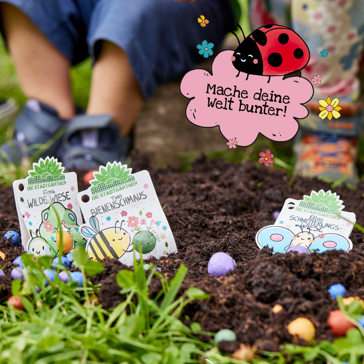 Zwei Kinder sitzen im Garten, im Vordergrund sind bunte Samenbomben und Pflanzschilder mit Illustrationen zu sehen; darüber eine Zeichnung eines Marienkäfers und der Text „Mache deine Welt bunter!“.