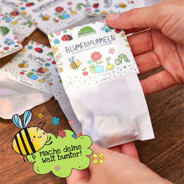 Eine Hand hält ein buntes Saatgut-Tütchen mit der Aufschrift "BLUMENMURMELN" über einem Holztisch; im Vordergrund ist eine gezeichnete Biene mit dem Text "Mache deine Welt bunter!".