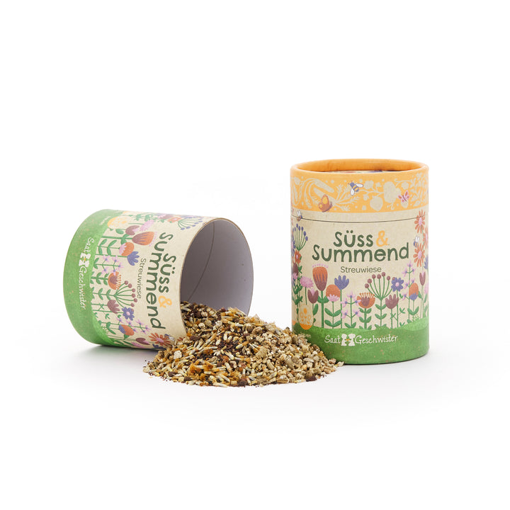 Zwei bunte Pappdosen mit Blumenmuster, eine stehend und eine liegend, aus der Blumensamen herausfallen, beschriftet mit „Süß & Summend Streuwiese“.