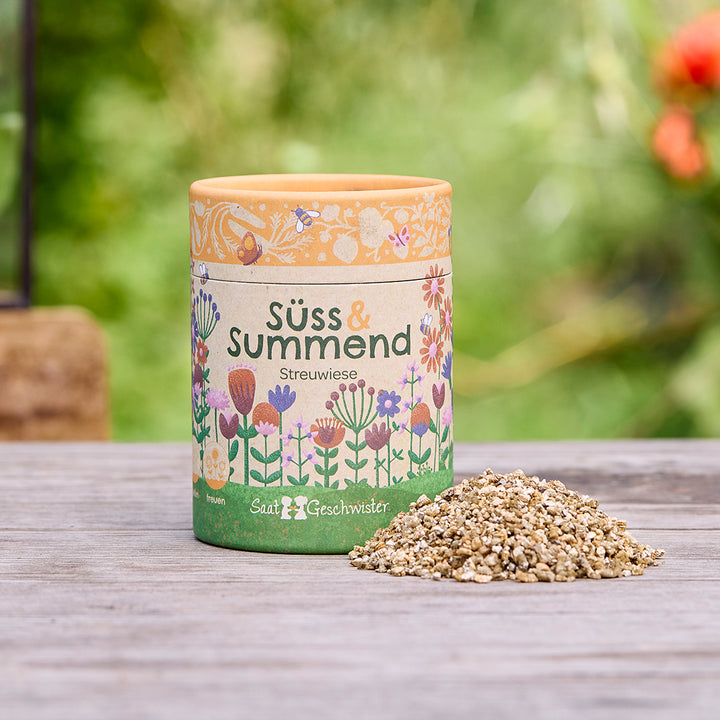 Runde Dose mit der Aufschrift „Süß & Summend Streuwiese“ und einem Blumenmotiv, daneben ein kleiner Haufen Saatgut auf einem Holztisch, unscharfer grüner Hintergrund.