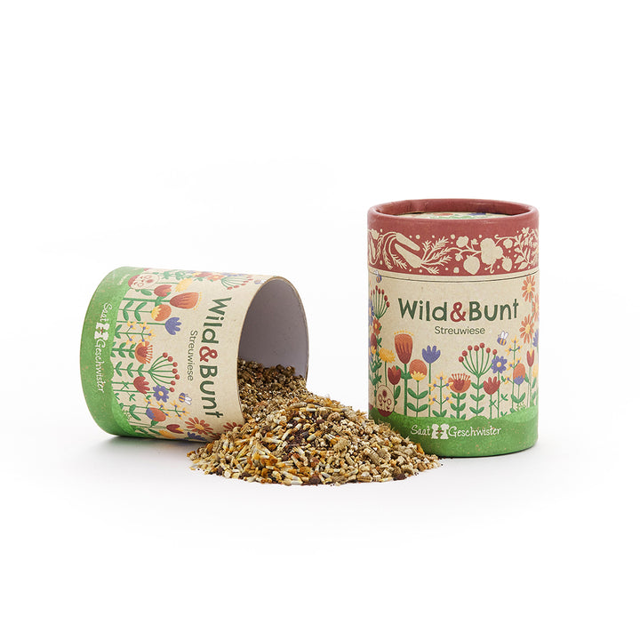 Zwei bunte Dosen mit der Aufschrift "Wild & Bunt Streuwiese", eine liegt und ist geöffnet, aus der Samen auf den Tisch fallen.
