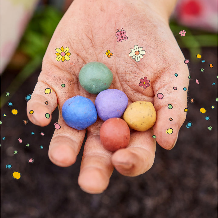 Eine Hand hält fünf bunte Samenbomben, umgeben von gezeichneten Blumen und bunten Punkten.