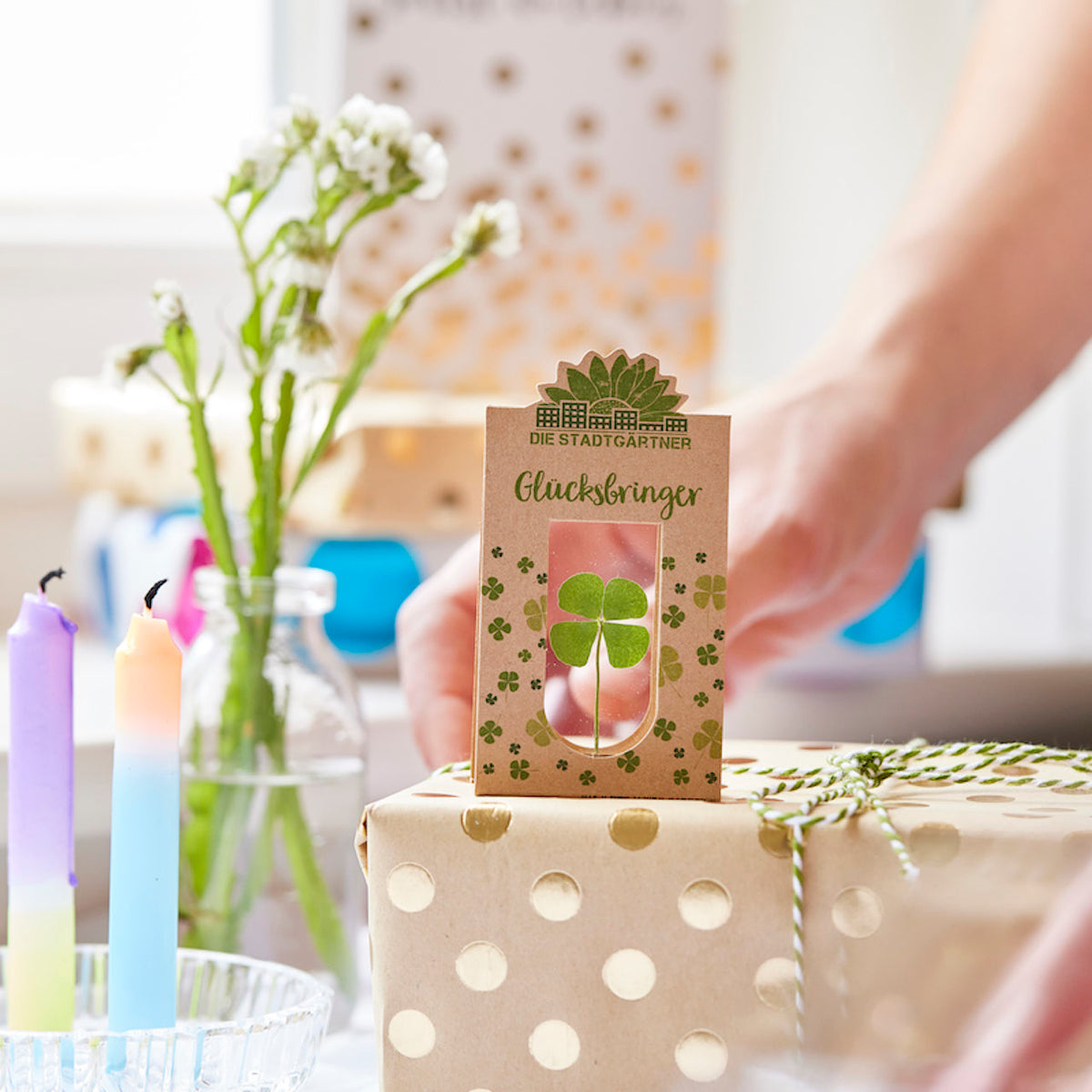 Eine Hand hält eine Verpackung mit einem vierblättrigen Kleeblatt und der Aufschrift "Glücksbringer" über einem Geschenk, im Hintergrund sind Blumen und bunte Kerzen zu sehen.