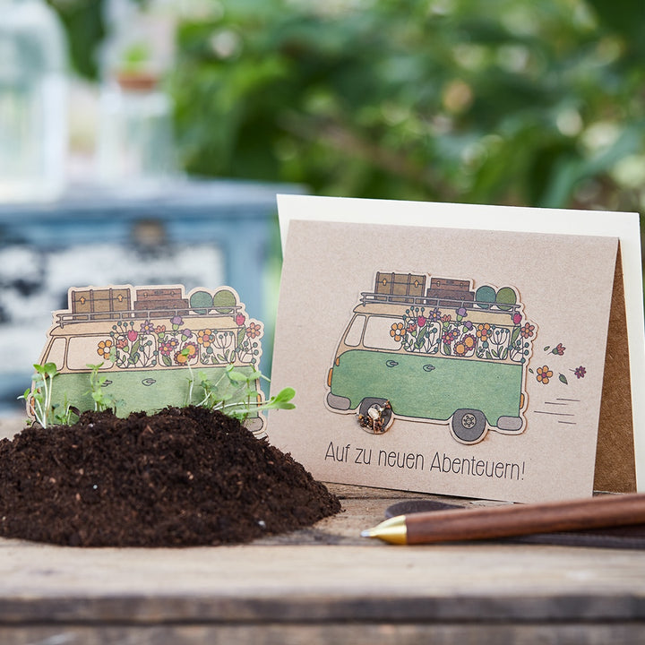 Grußkarte mit gezeichnetem grünen Bus voller Blumen und Gepäck, daneben Erde mit kleinen Pflänzchen, Text auf der Karte: „Auf zu neuen Abenteuern!“.