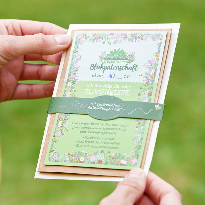 Person hält eine Karte mit Aufschrift "Blühpateenschaft" und Beschreibung eines Geschenks in Form einer Blumenwiese.