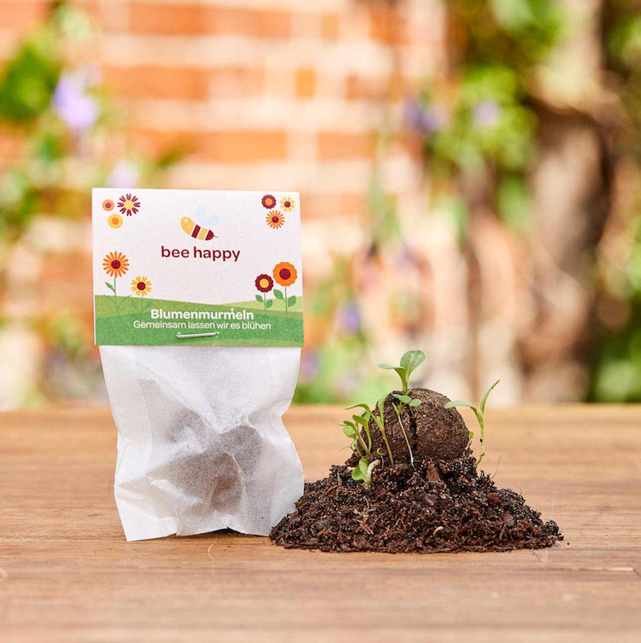 Blumenmurmeln-Packung mit der Aufschrift "bee happy" und kleinen Pflanzen, die aus einer Samenbombe in Erde sprießen, vor unscharfem Gartenhintergrund.