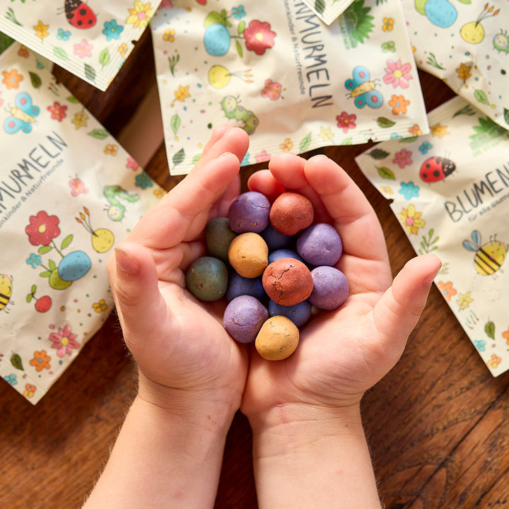 Kinderhände halten bunte Samenbomben, im Hintergrund liegen Tütchen mit Blumenmotiven auf einem Holztisch.