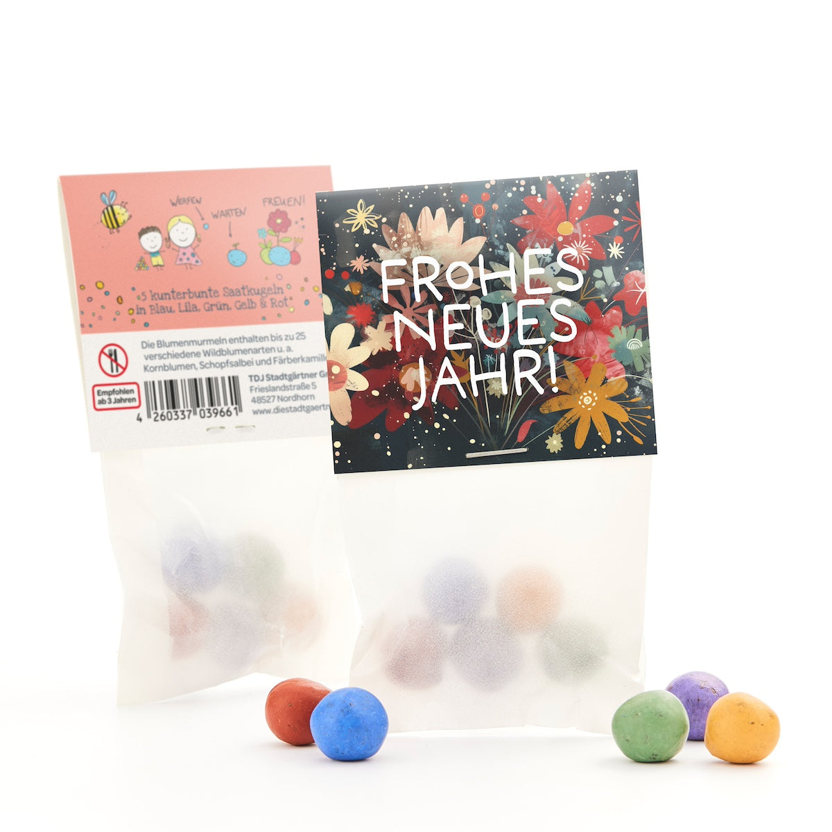 Zwei Tüten mit bunten Saatkugeln, eine mit blumigem "Frohes neues Jahr!"-Aufdruck und eine mit Beschreibung und Zeichnungen, einige Kugeln liegen davor.