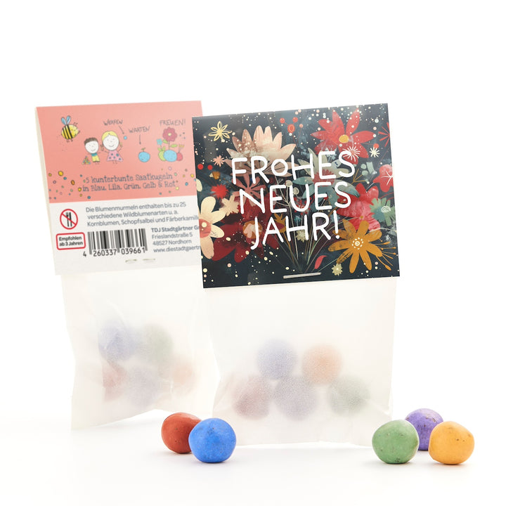 Zwei Tüten mit bunten Saatkugeln, eine mit blumigem "Frohes neues Jahr!"-Aufdruck und eine mit Beschreibung und Zeichnungen, einige Kugeln liegen davor.
