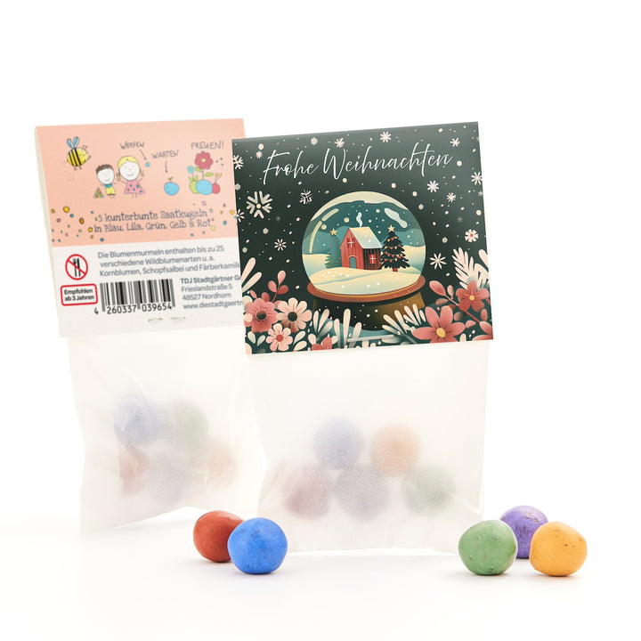 Zwei Tütchen mit bunten Saatkugeln, eines mit einer weihnachtlichen Grußkarte mit Schneekugelmotiv und Aufschrift „Frohe Weihnachten“, daneben bunte Kugeln auf weißem Hintergrund.