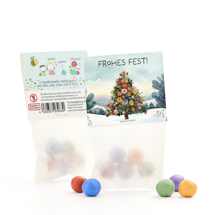 Zwei Tütchen mit bunten Saatkugeln, eine mit Weihnachtsbaum und "Frohes Fest!"-Gruß, verschiedene bunte Kugeln davor.
