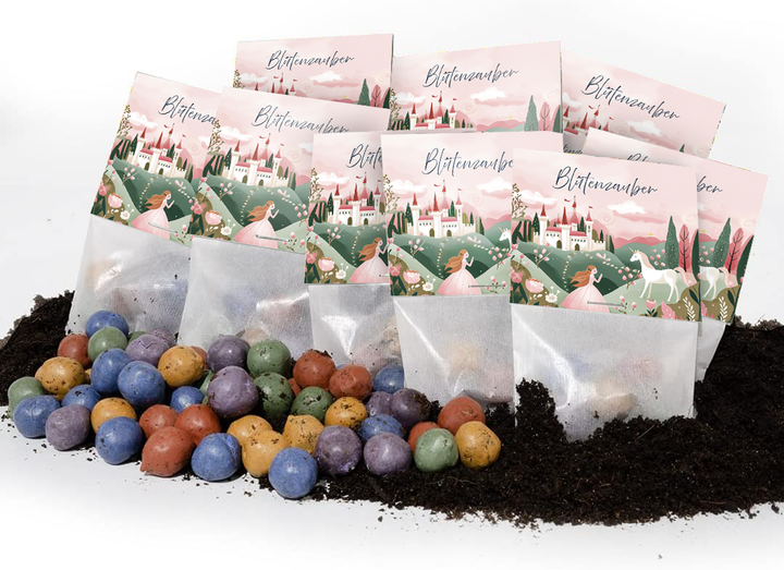 Mehrere Tüten mit der Aufschrift „Blütenzauber“ und märchenhaftem Design, davor bunte Samenbomben auf Erde liegend.