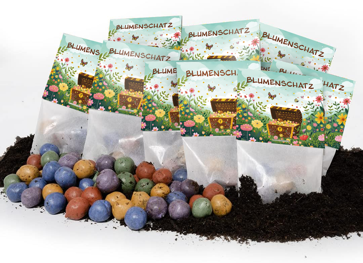 Mehrere bunte Samenbomben liegen vor aufrecht stehenden Tütchen mit der Aufschrift "Blumenschatz" und einer Illustration von Blumen, Schmetterlingen und einer Schatztruhe auf Erde.